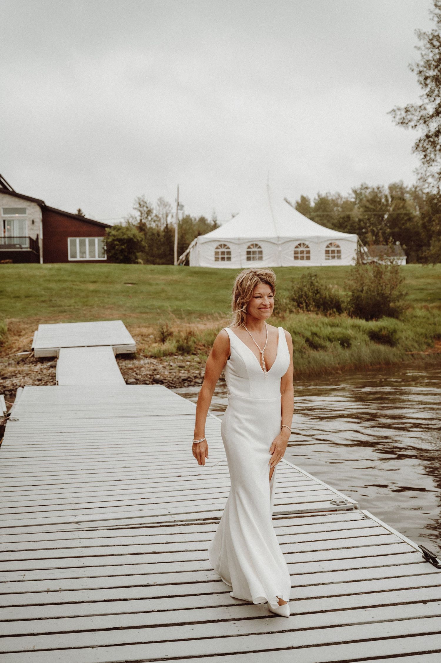Bride in elegant white dress poses on wooden dock with white tent and lake in background.