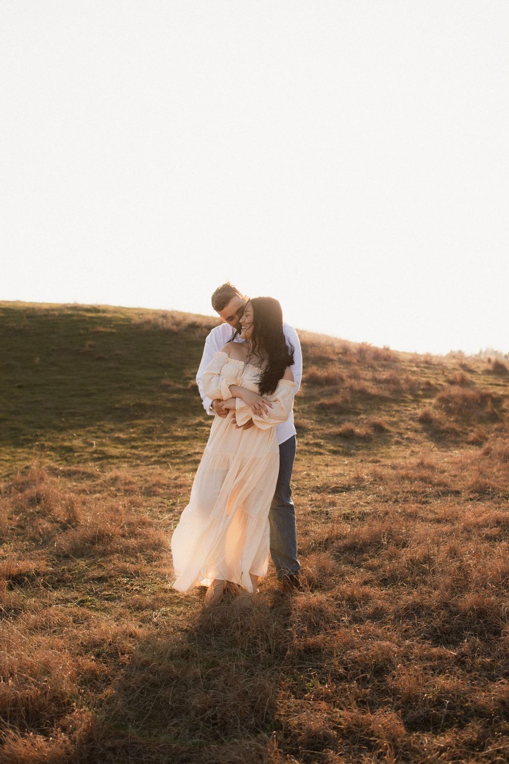 A romantic couple embraces on a hillside during golden hour sunset while wearing white and formal attire.