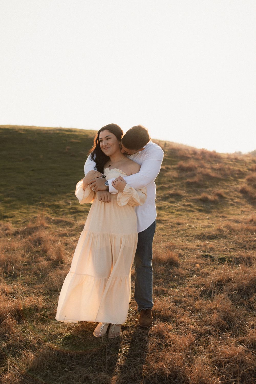 A couple shares an intimate moment on a grassy hillside at sunset, with her wearing a flowing cream dress.