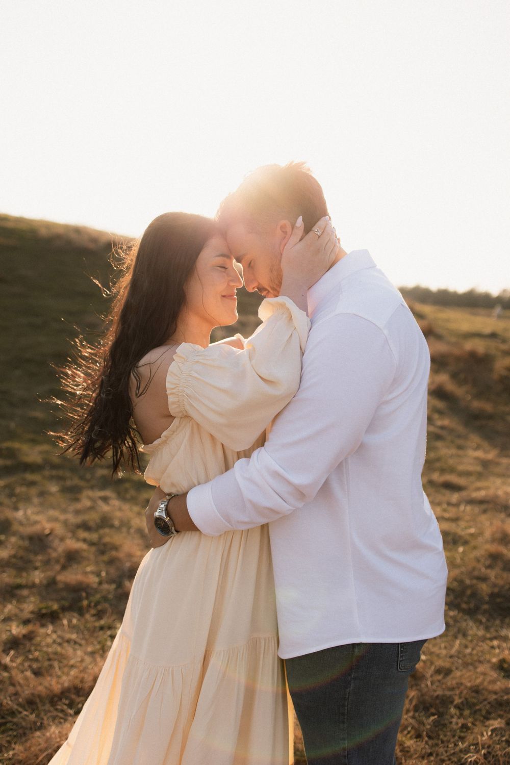 A romantic couple embraces in a sunlit field at golden hour, with her cream dress flowing in the breeze.