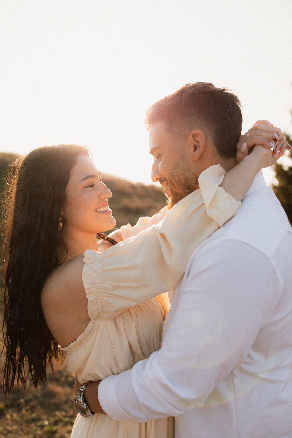 A couple embraces in warm sunset light, with her in a cream dress and him in a white shirt enjoying an intimate moment outdoors.