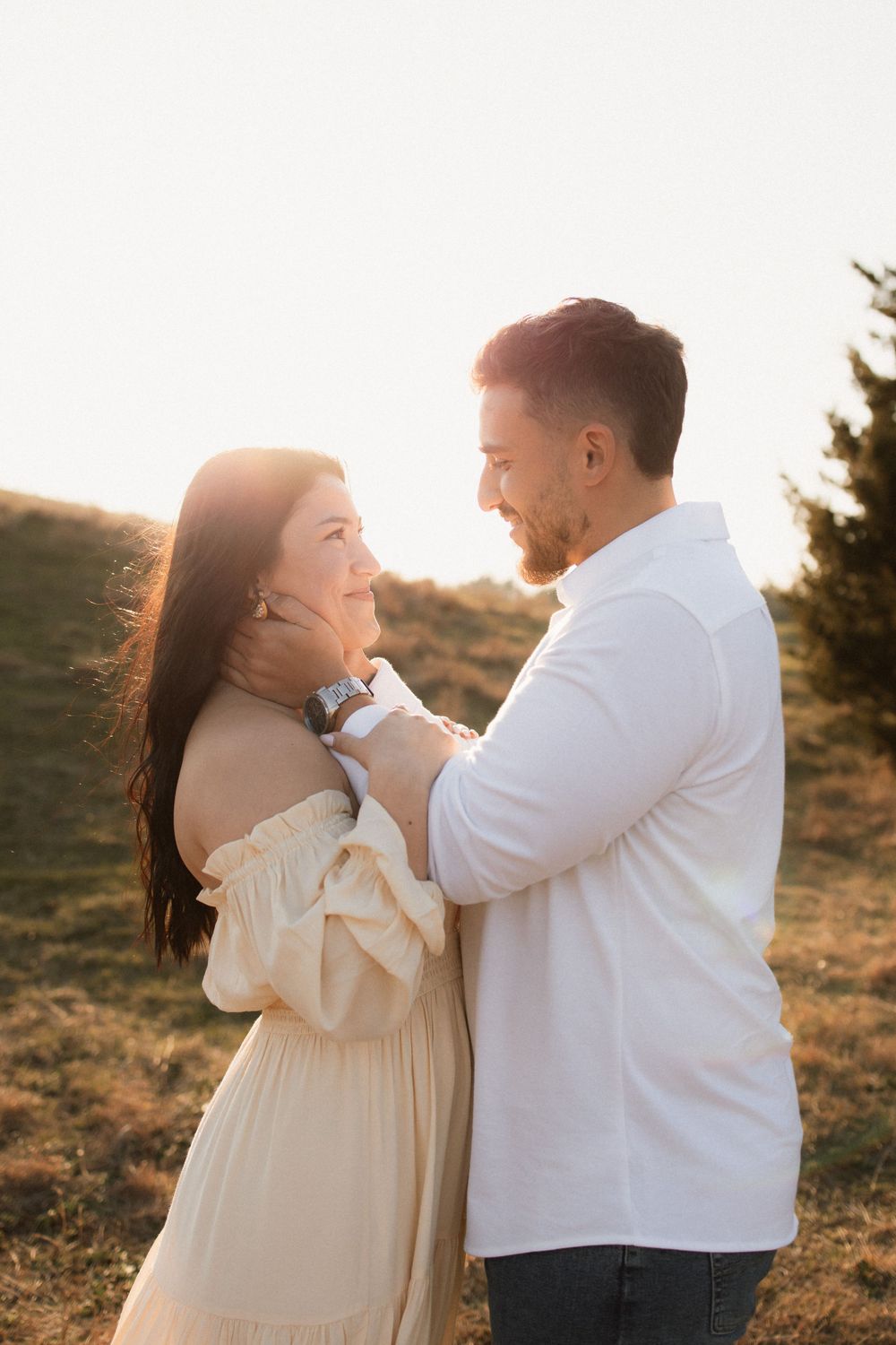 Romantic couple shares tender moment at sunset on a grassy hillside with woman in cream dress and man in white shirt.