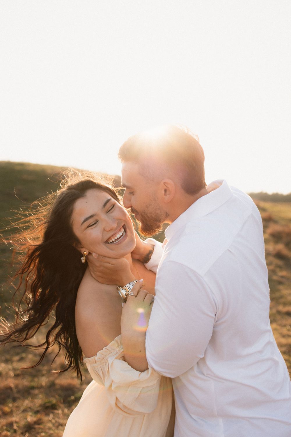 A joyful couple laughs together on a hill during sunset in matching white outfits.
