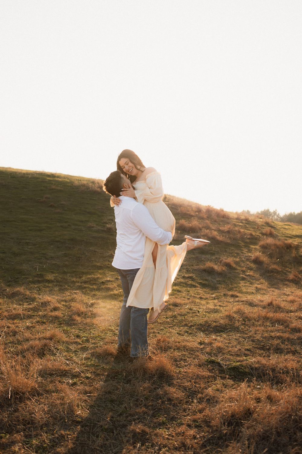 A romantic couple embraces on a grassy hillside during golden hour, with one person lifting the other.