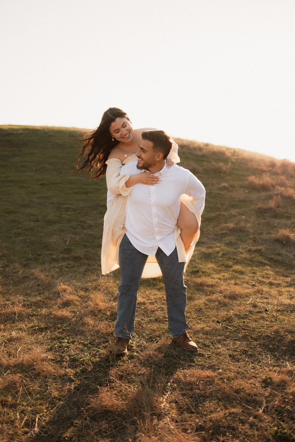 A playful moment captured on a grassy hillside during sunset as a couple share a piggyback ride.
