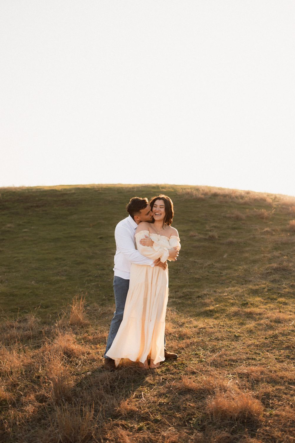 A couple embraces on a grassy hillside during golden hour sunset, with soft lighting illuminating their romantic moment.