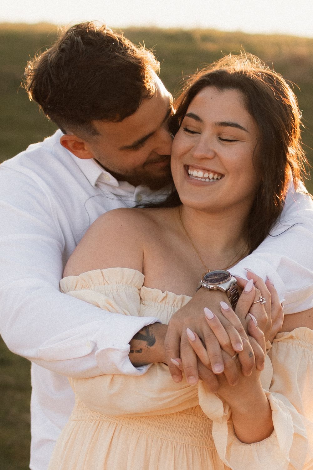 Romantic couple embraces in warm sunset light while sharing intimate moment in off-shoulder dress and white shirt.