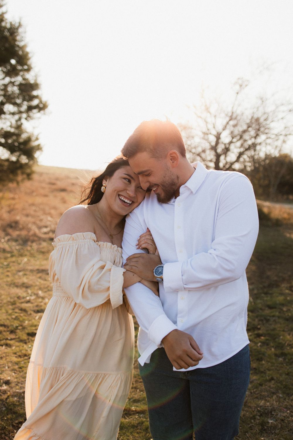 Couple sharing a romantic laugh while embracing in golden sunlight on a grassy hillside.