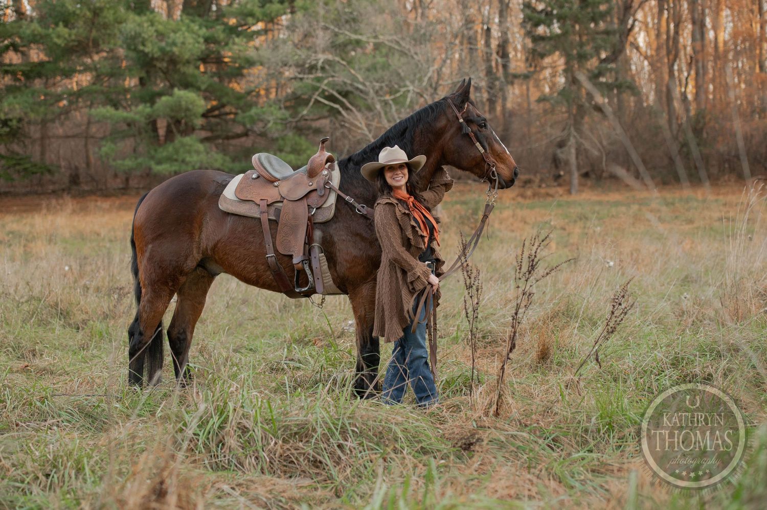 A western rider stands beside a saddled brown horse in a rustic autumn field at sunset.