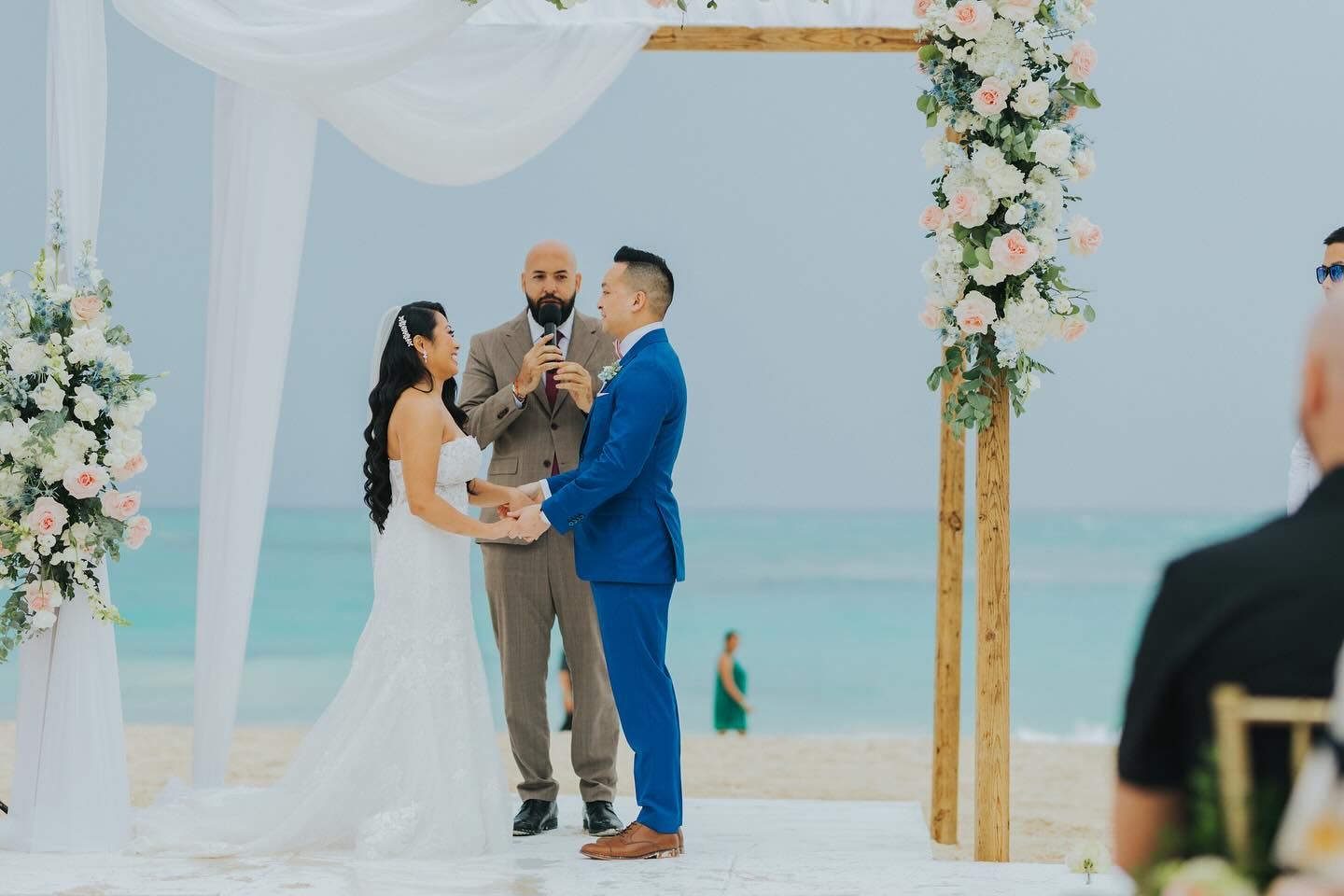 A beachfront wedding ceremony under a wooden arch decorated with white flowers and flowing fabric beside turquoise waters.