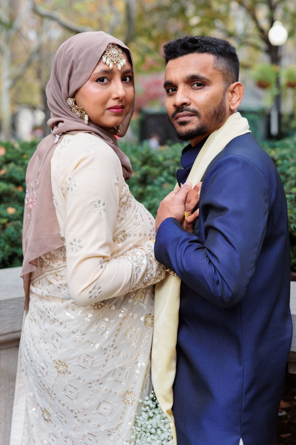 Muslim bride in hijab and gold jewelry posing with groom in blue sherwani outdoors.
