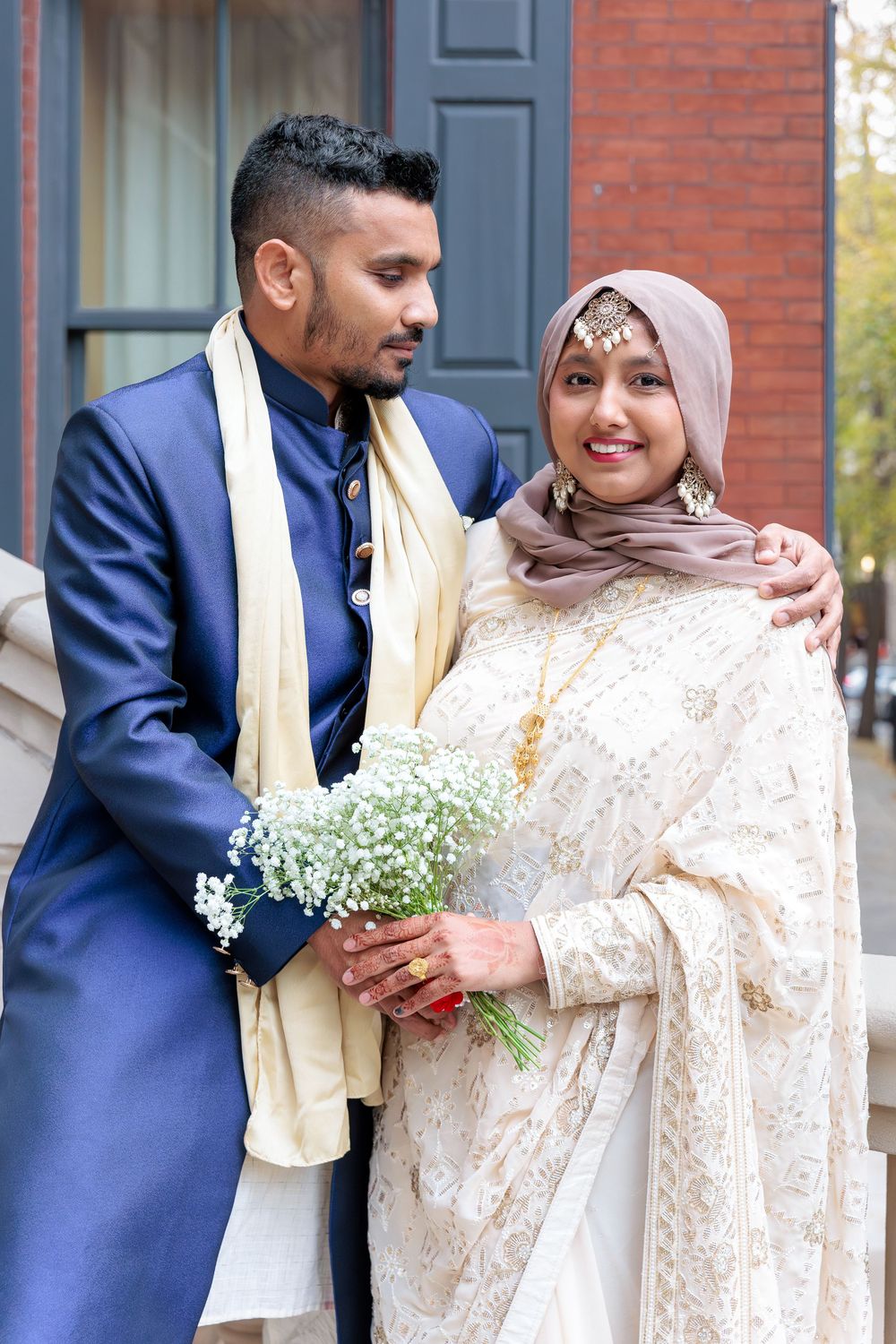 South Asian groom in blue sherwani embracing bride in hijab holding white flower bouquet.