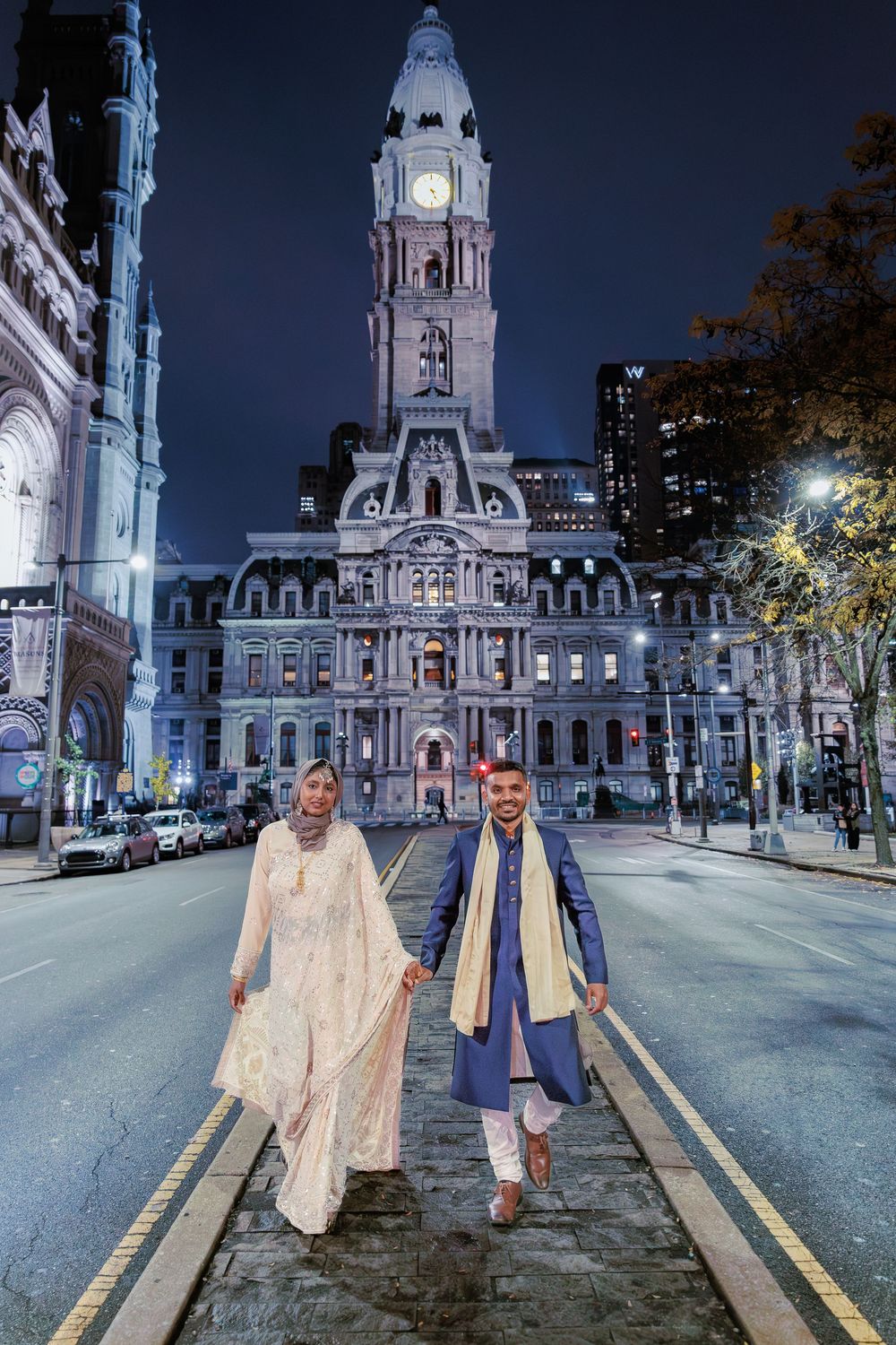 Muslim couple in traditional wedding attire walking hand in hand with Philadelphia City Hall lit up behind them.