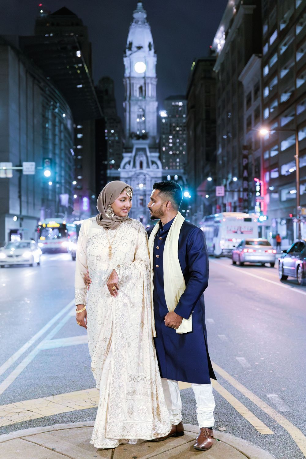 Muslim bride in hijab and groom in blue sherwani posing on a city street at night.