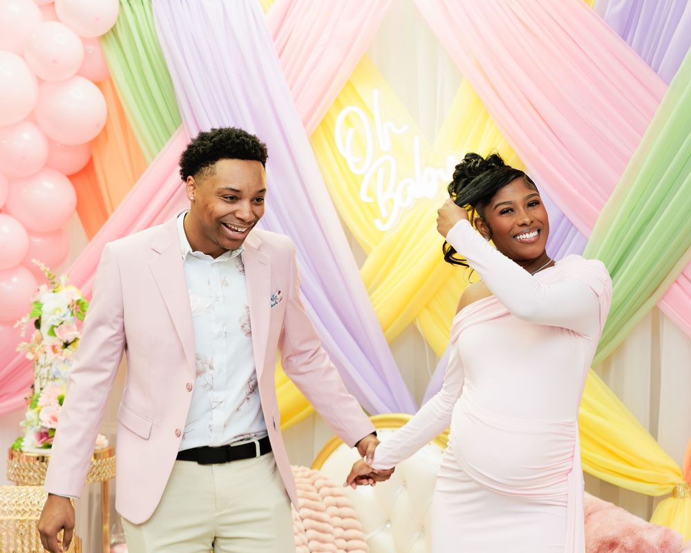 Joyful couple laughing and holding hands at a colorful pastel balloon baby shower celebration.
