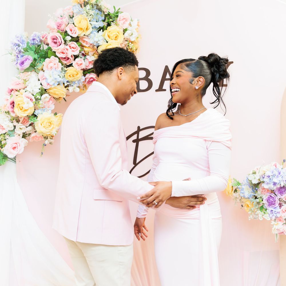 Expecting couple sharing a tender moment at a floral baby shower backdrop, woman in white maternity gown.