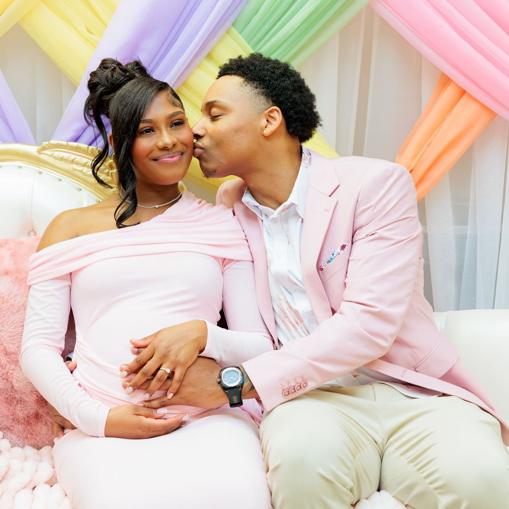 Couple at a pastel-themed baby shower, woman in pink maternity dress receiving a kiss while holding her baby bump.