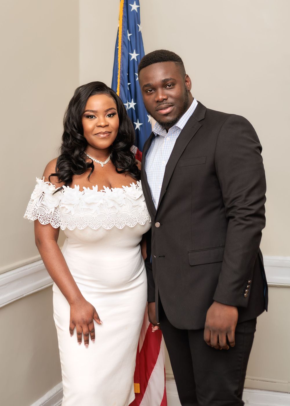 Bride in white off-shoulder lace dress and groom in dark suit smiling together beside an American flag.