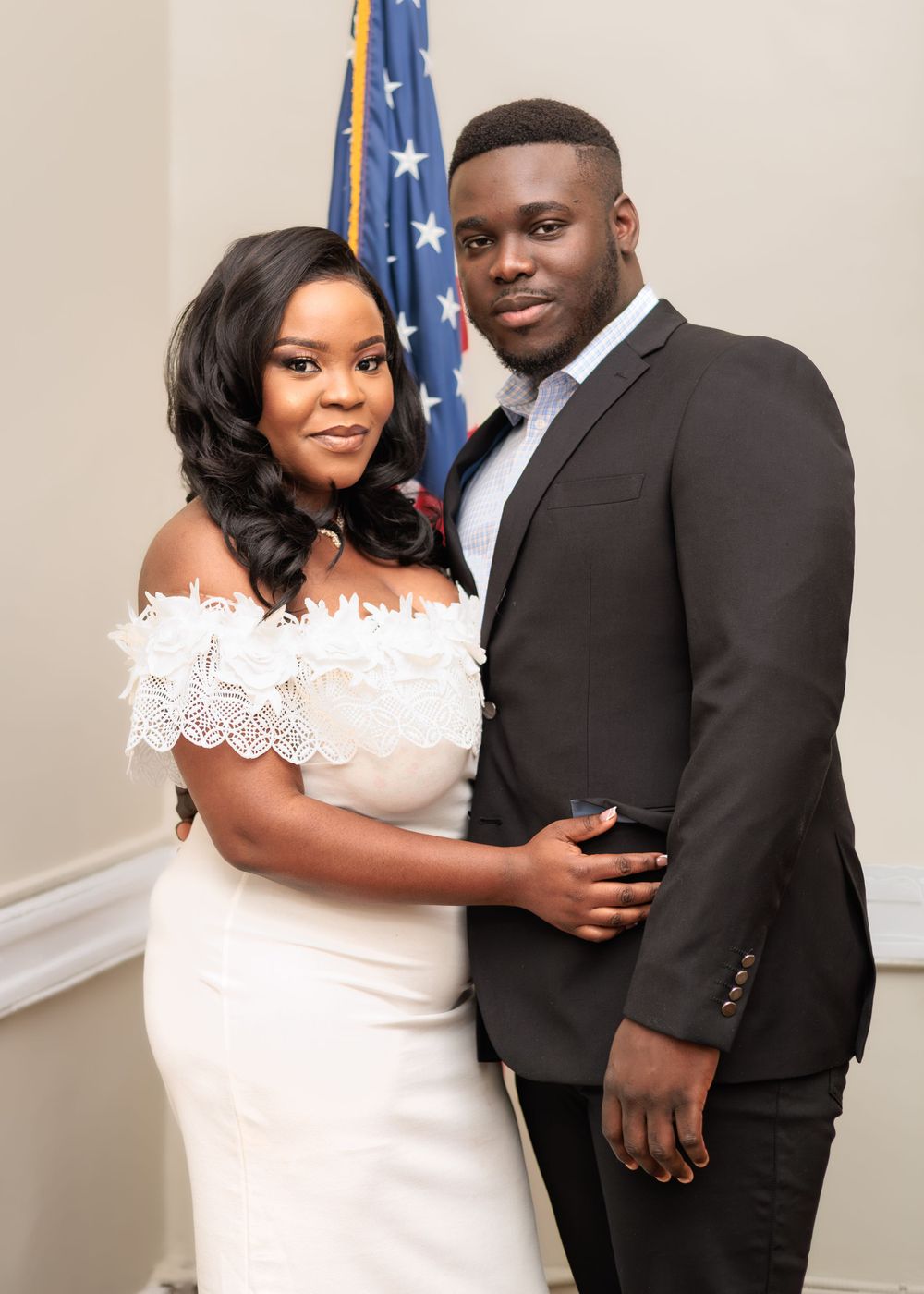 Couple posing elegantly, woman in white lace off-shoulder dress and man in dark suit, with American flag backdrop.
