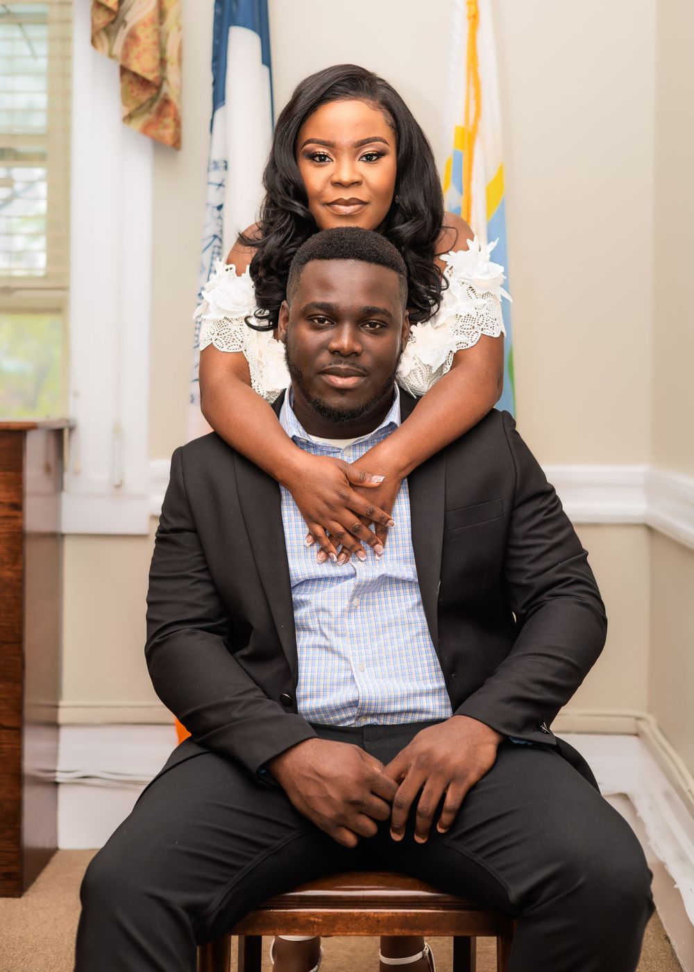 Elegant Black couple poses formally indoors, woman stands behind seated man in a black suit.