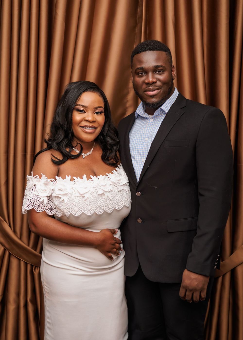 An elegant couple poses together before a golden curtain backdrop, woman in white off-shoulder dress.
