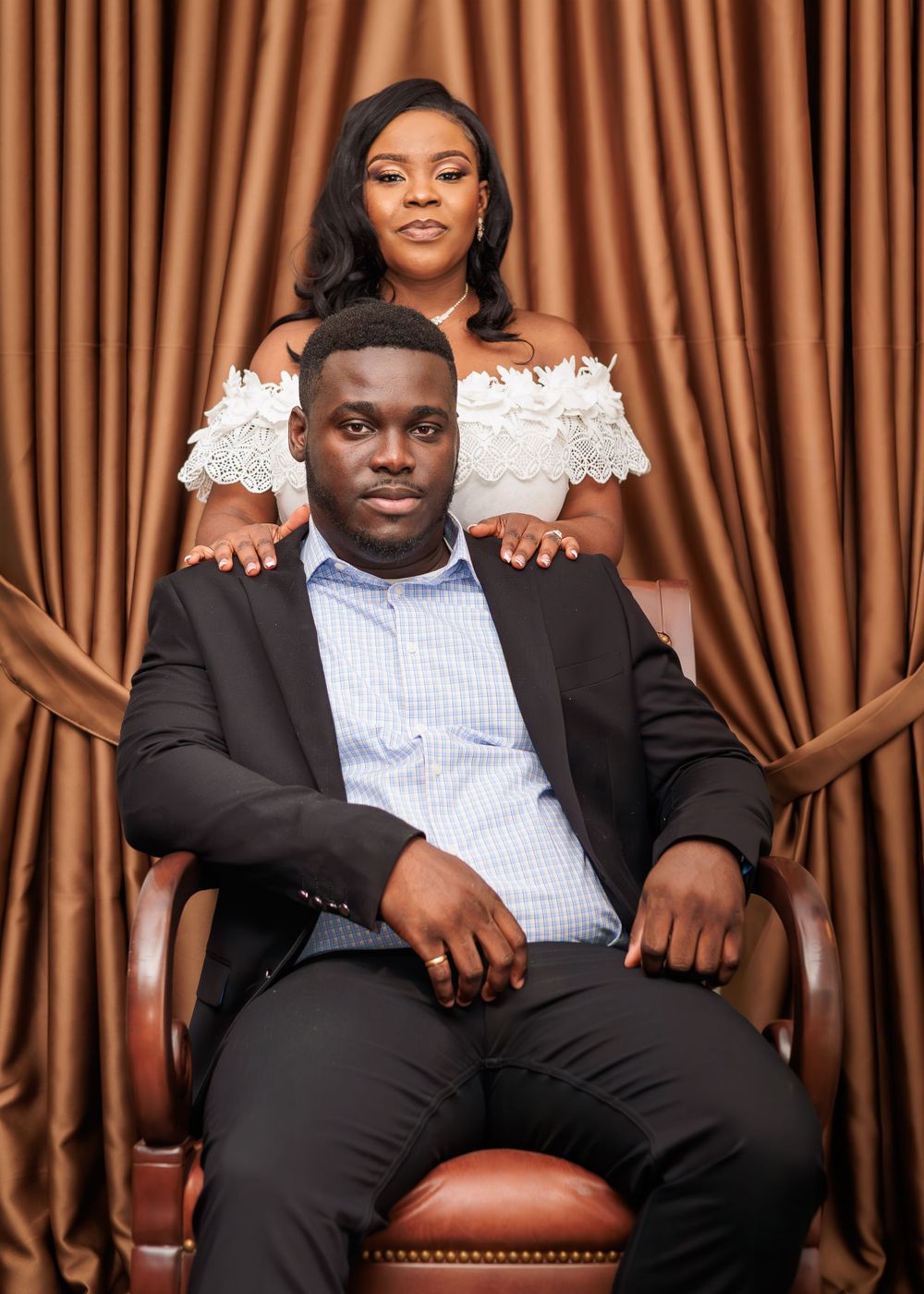 Groom seated in leather chair with bride standing behind him, posing for formal portrait against draped curtains.