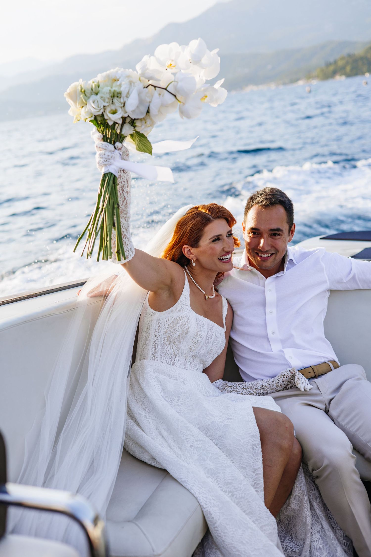 A couple in white attire share a romantic moment on a boat with mountains and blue water in the background.