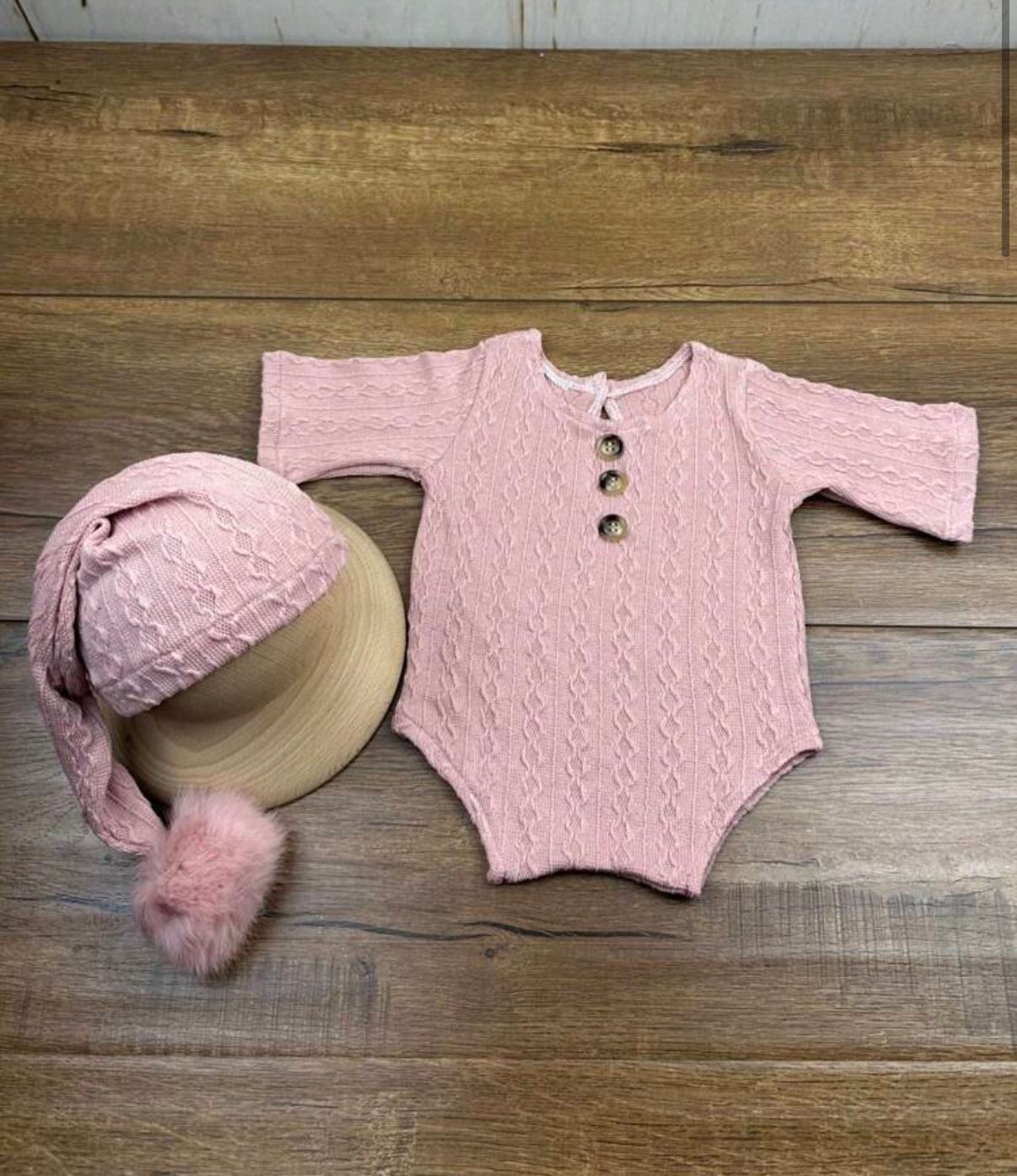 Pink knitted baby romper and hat set laid flat on wooden background.