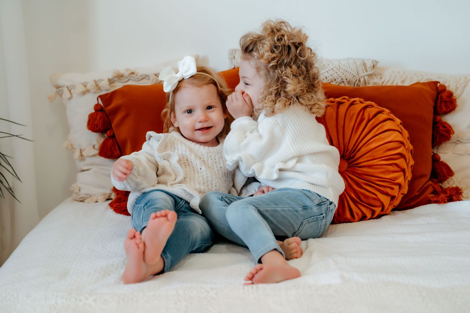 Two young children in casual attire sit together on a couch with orange throw pillows in a cozy home setting.