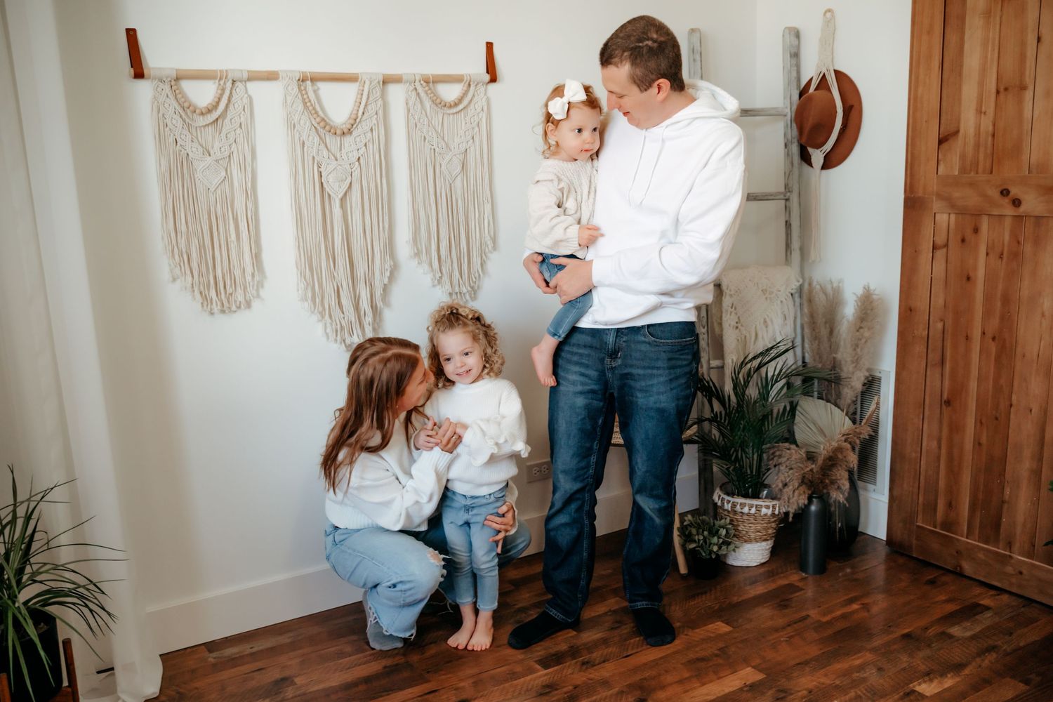 Cozy interior scene with boho wall decor, wooden floors, and a family sharing a sweet moment near potted plants.