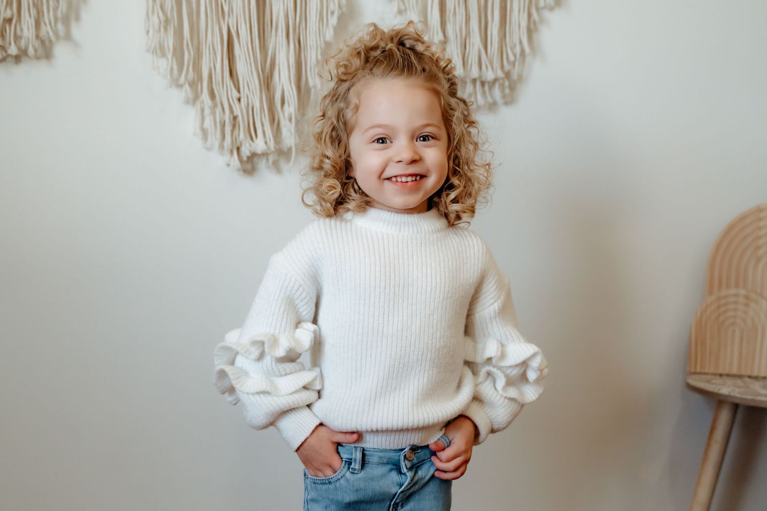 A child wearing a cozy white knit sweater with balloon sleeves and jeans poses against a neutral wall with macrame decor.