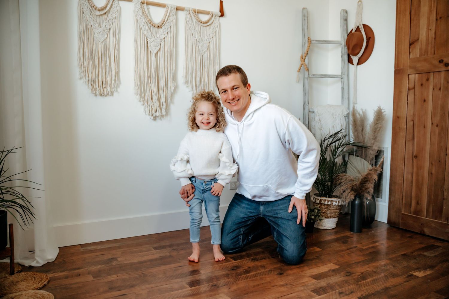 A casual indoor portrait against a wall decorated with macrame hangings and featuring hardwood flooring and rustic decor.