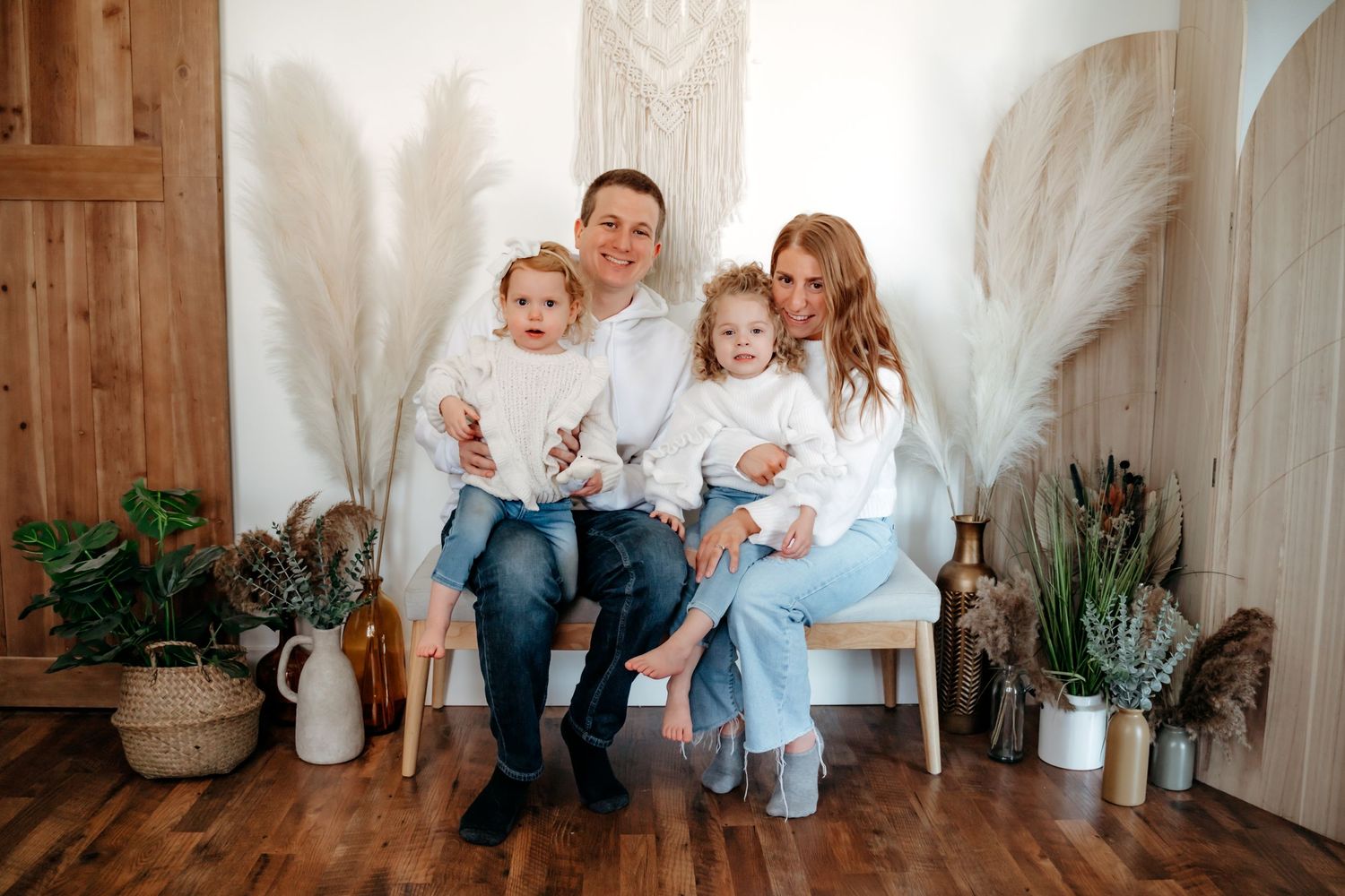 Family wearing white and blue sits together on a white bench surrounded by potted plants in a rustic room.