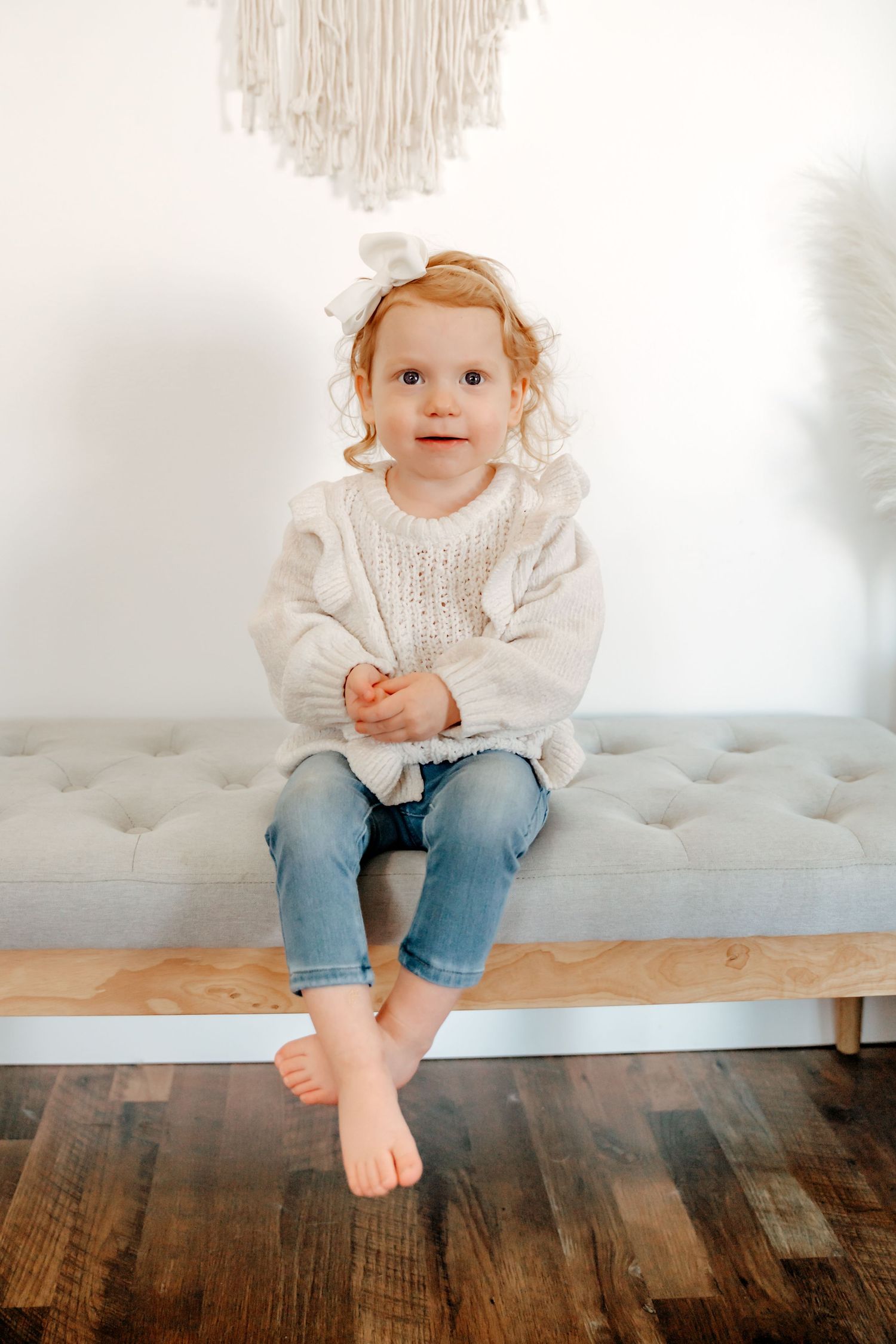 A toddler in a white sweater and jeans sits on a gray tufted bench with bare feet dangling.