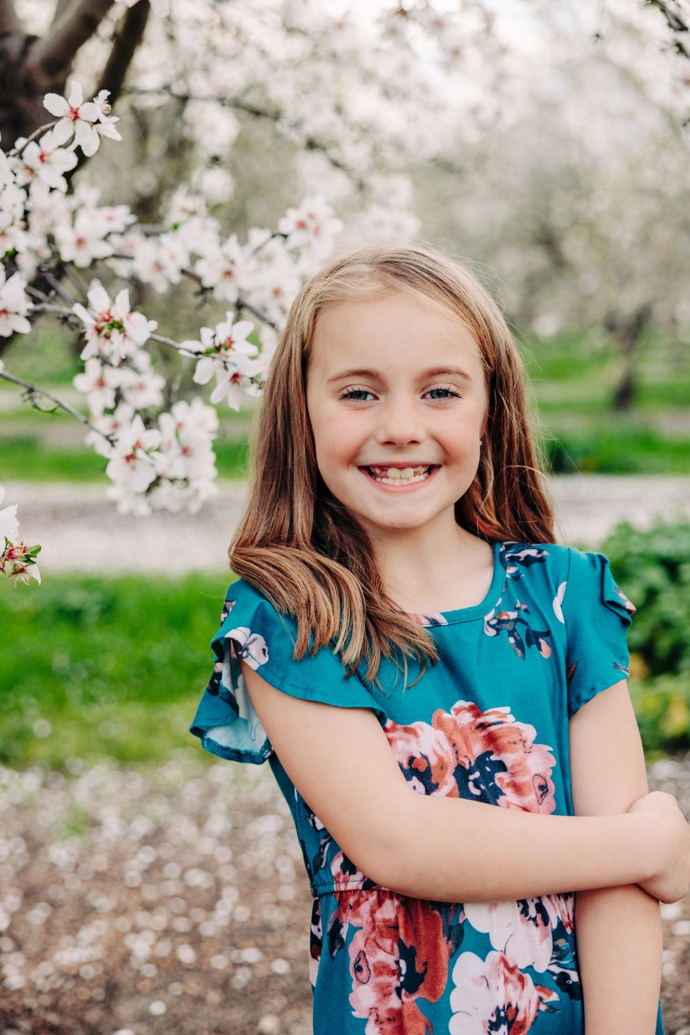 A child in a blue floral dress smiles while standing near blooming cherry blossom trees in springtime.