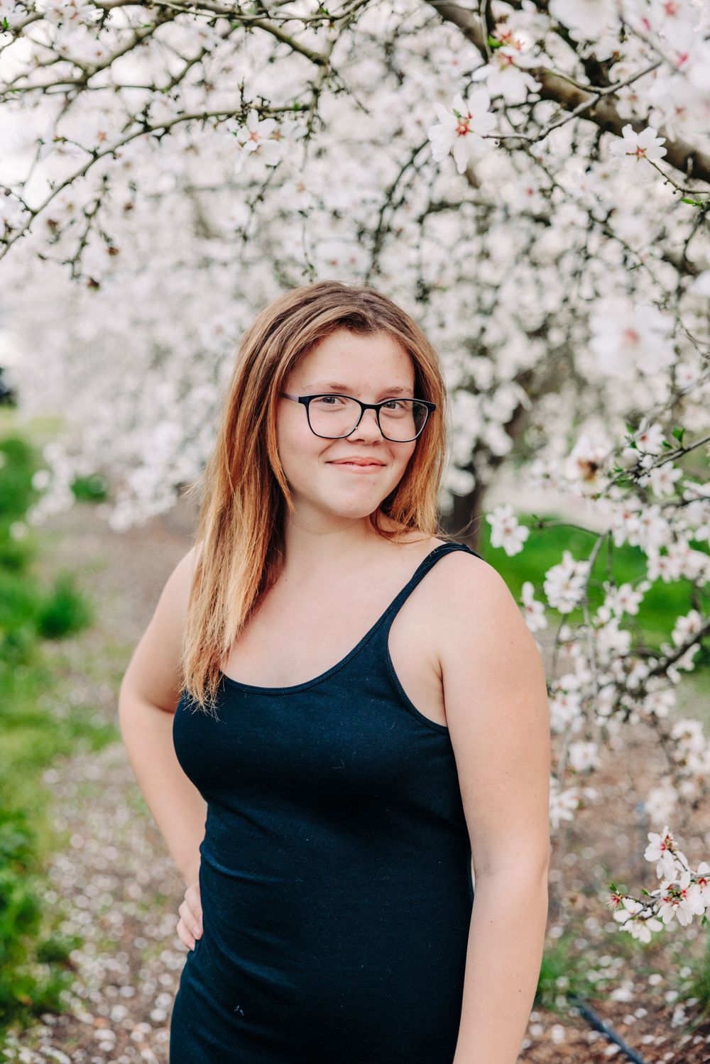A person in a black tank top poses among blooming white cherry blossoms on a spring day.