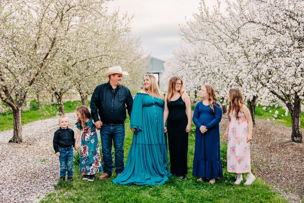 A group stands together in a blooming cherry orchard with white blossoms on a spring day, wearing coordinated blue dresses.