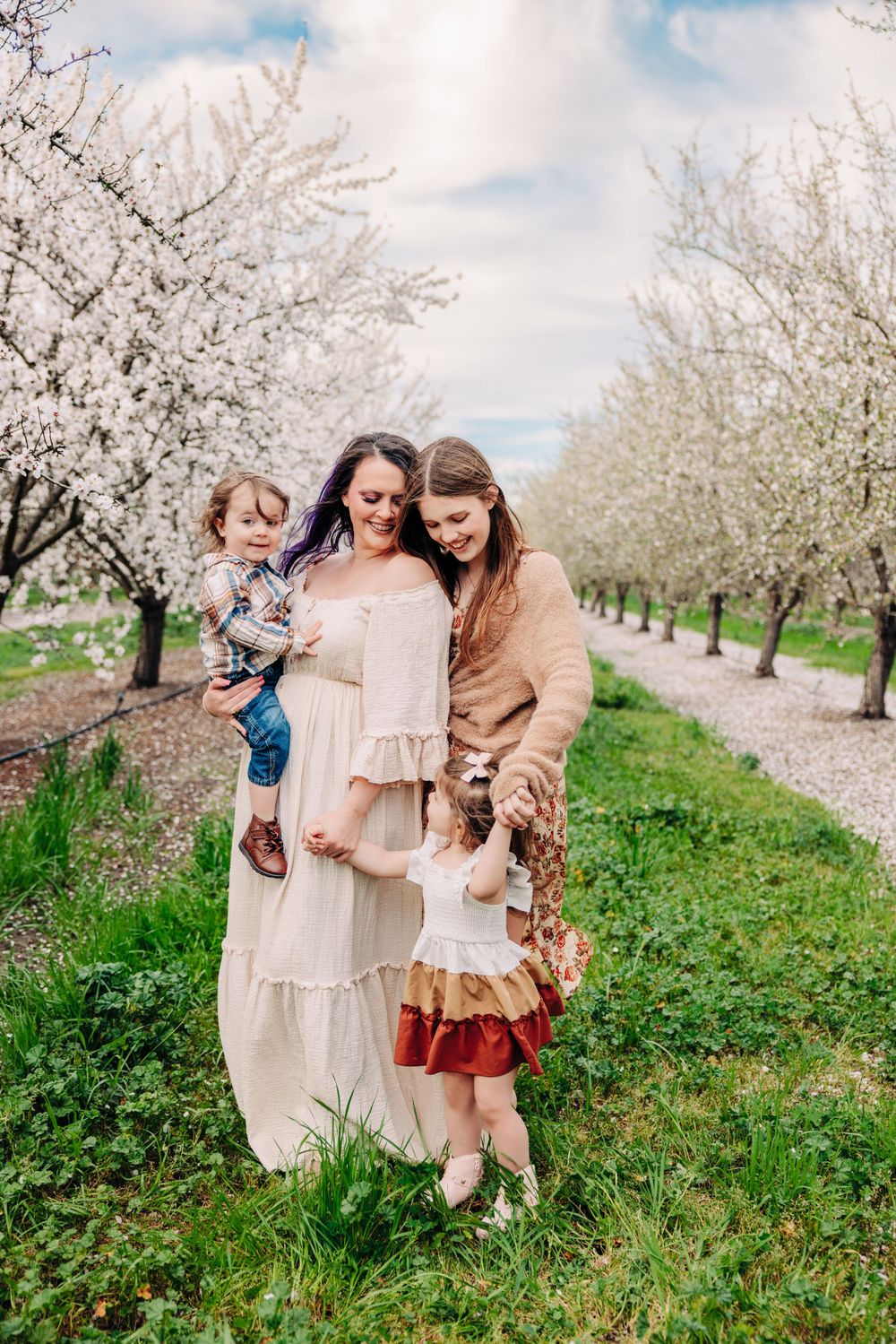 People in coordinated neutral outfits standing between rows of blooming white cherry blossom trees in an orchard.
