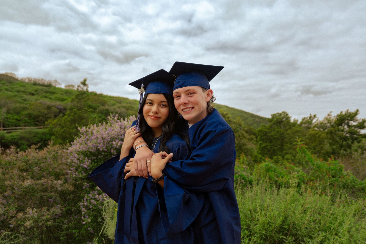 Two graduates wearing blue caps and gowns embrace while celebrating outdoors on a cloudy day with green hills in background.