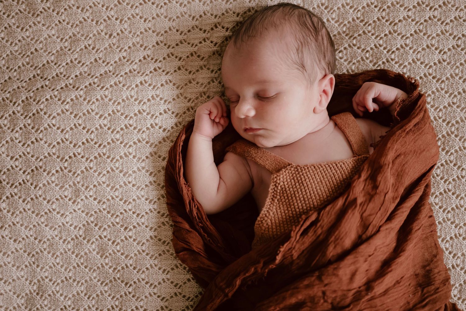 Newborn baby sleeping peacefully wrapped in brown blanket on textured beige background.