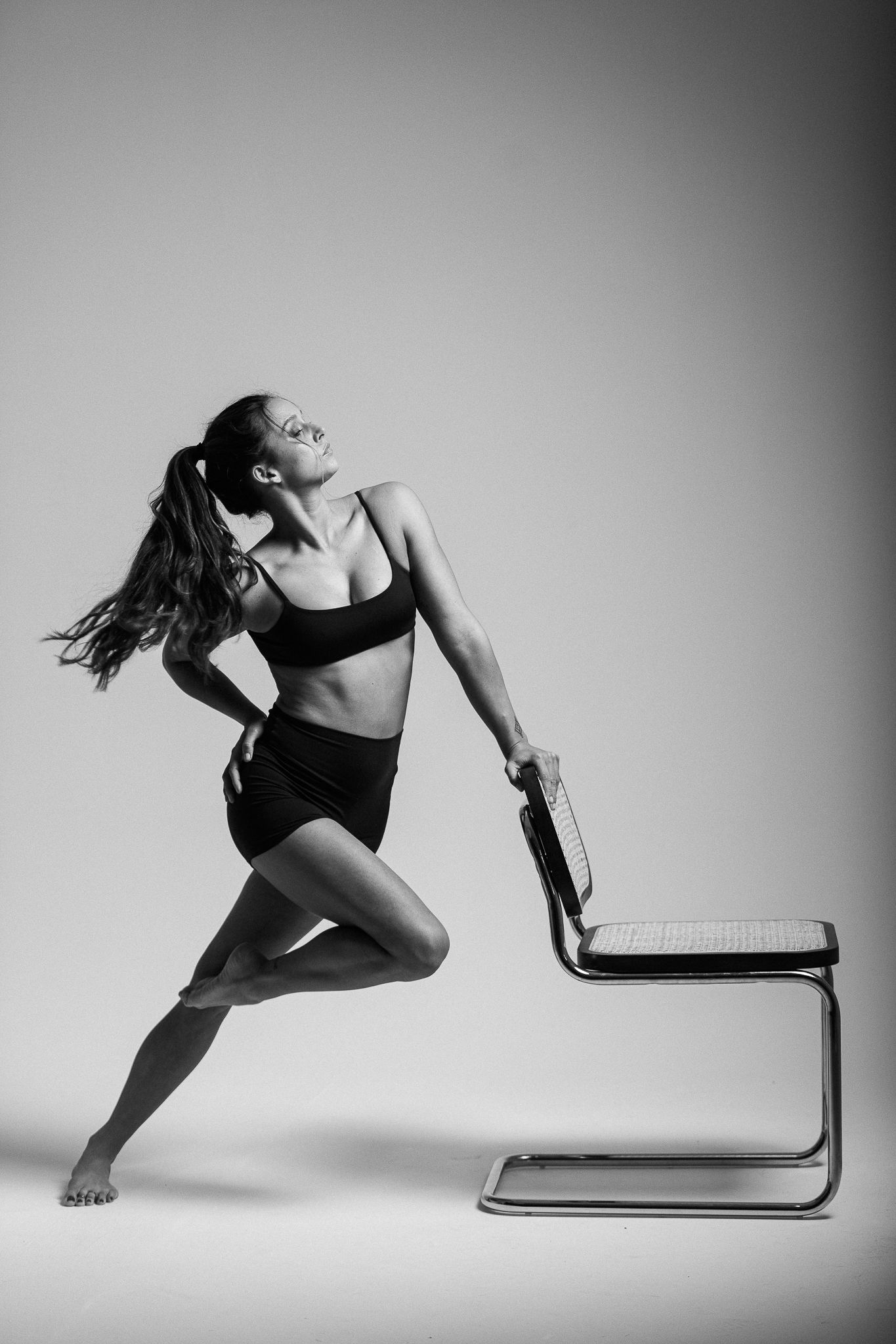 A dancer in black athletic wear strikes a dynamic pose while using a minimalist chair against a grayscale background.