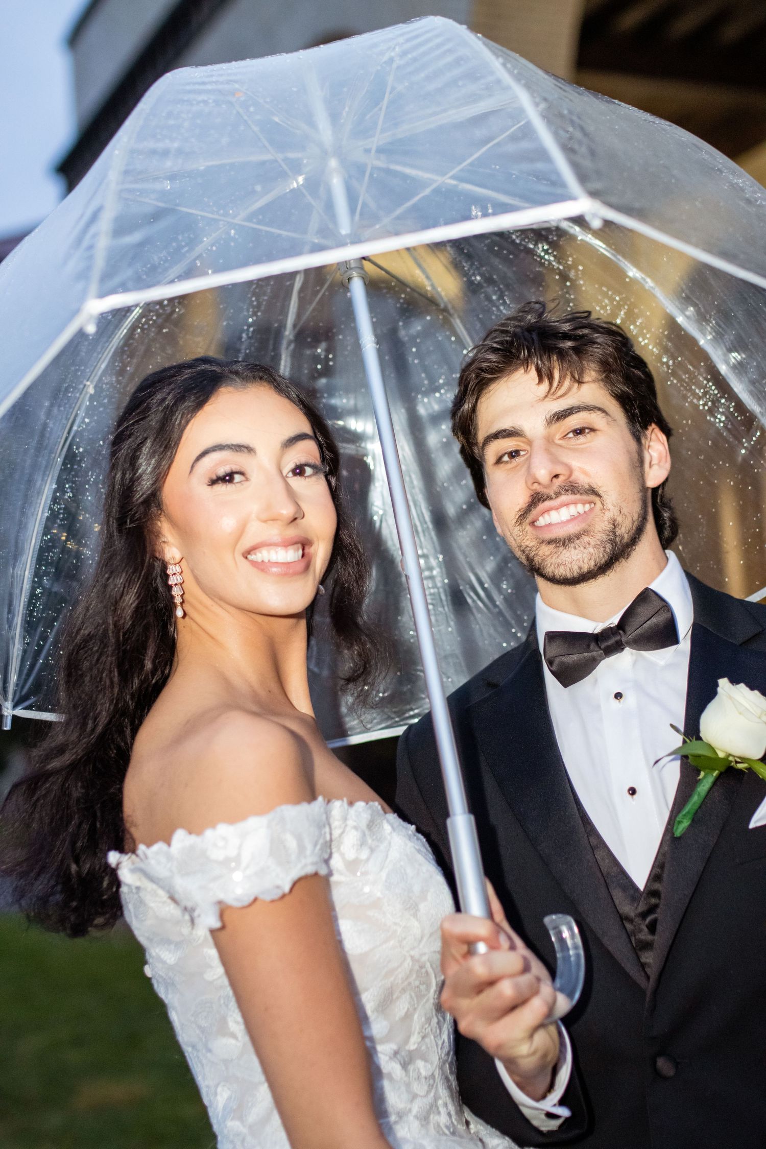A bride and groom smile while sharing a clear umbrella, with the bride wearing an off-shoulder lace gown.