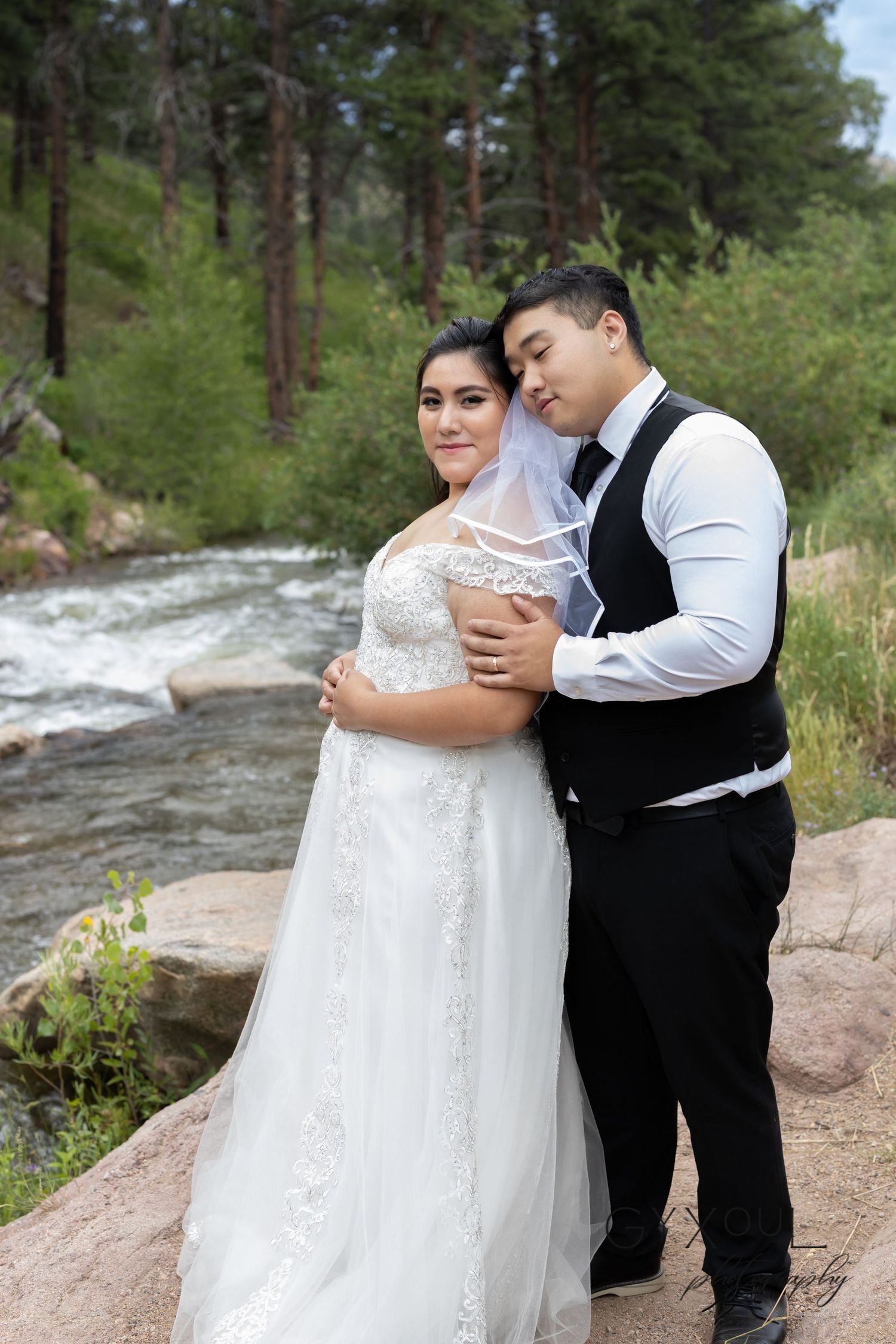 Newlyweds embrace on rocky terrain beside a rushing stream in a forest setting.