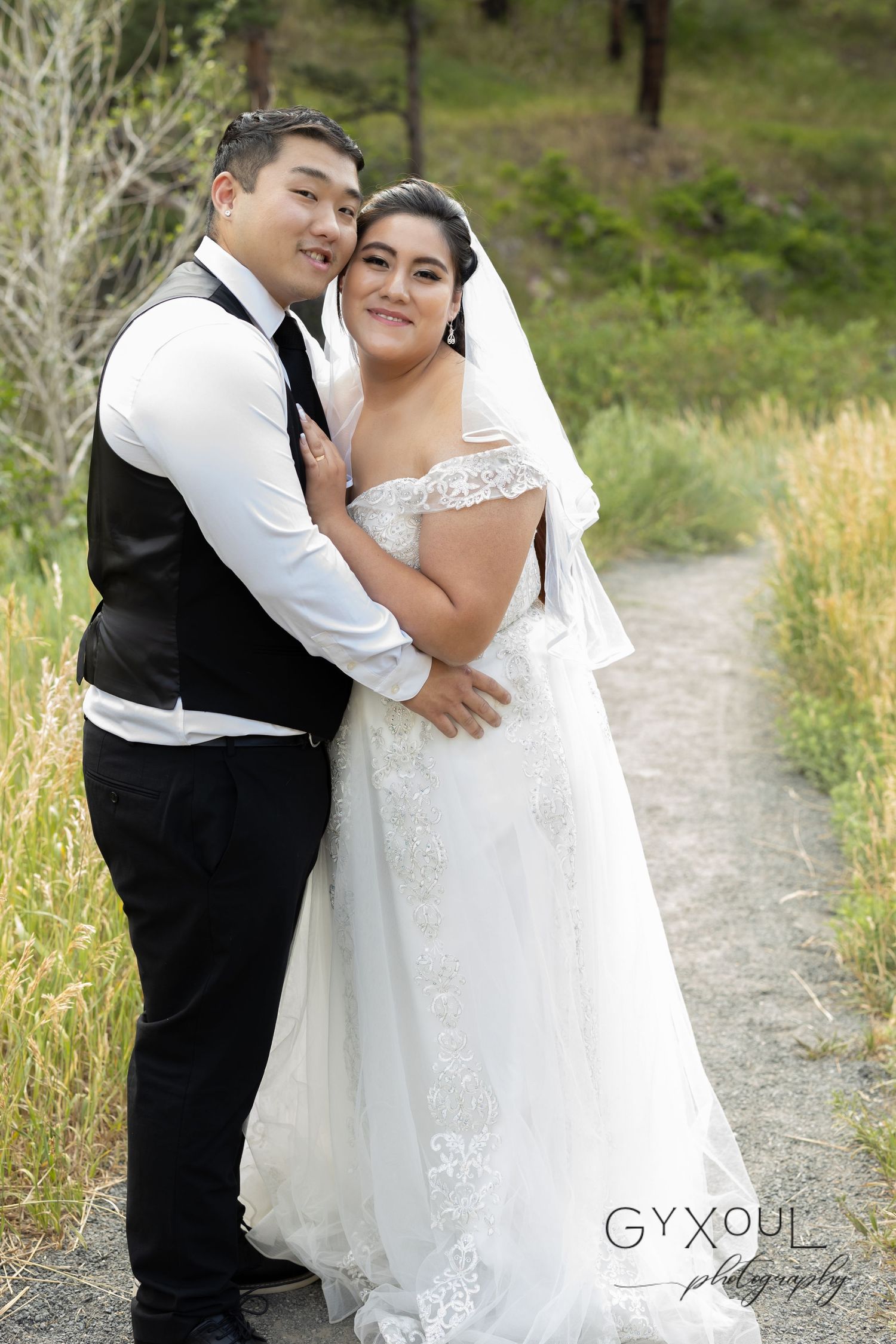 Happy newlyweds embrace on a nature path surrounded by greenery and trees.