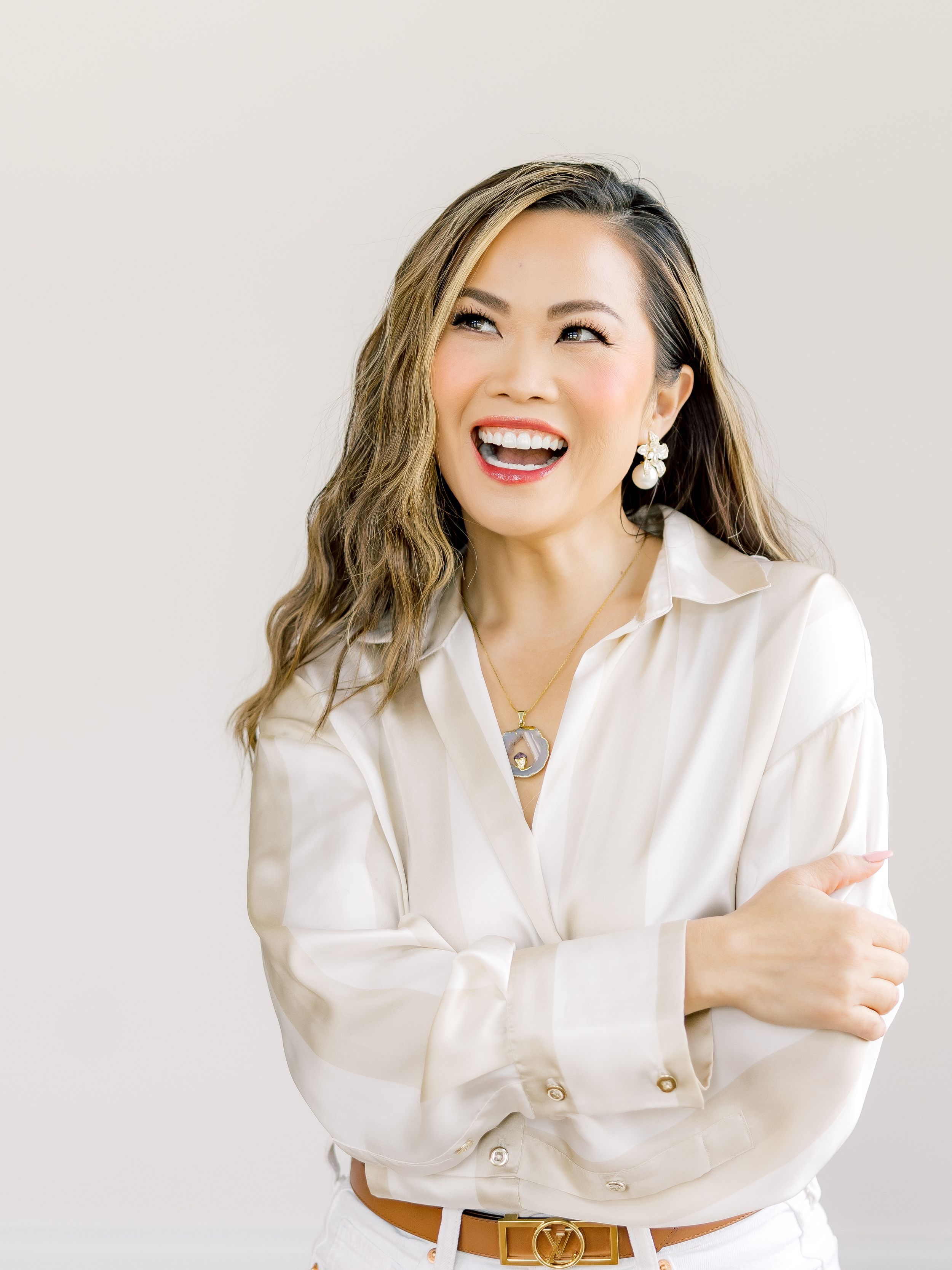 Woman in white button-down shirt laughs joyfully while wearing dangling earrings and necklace against neutral background.