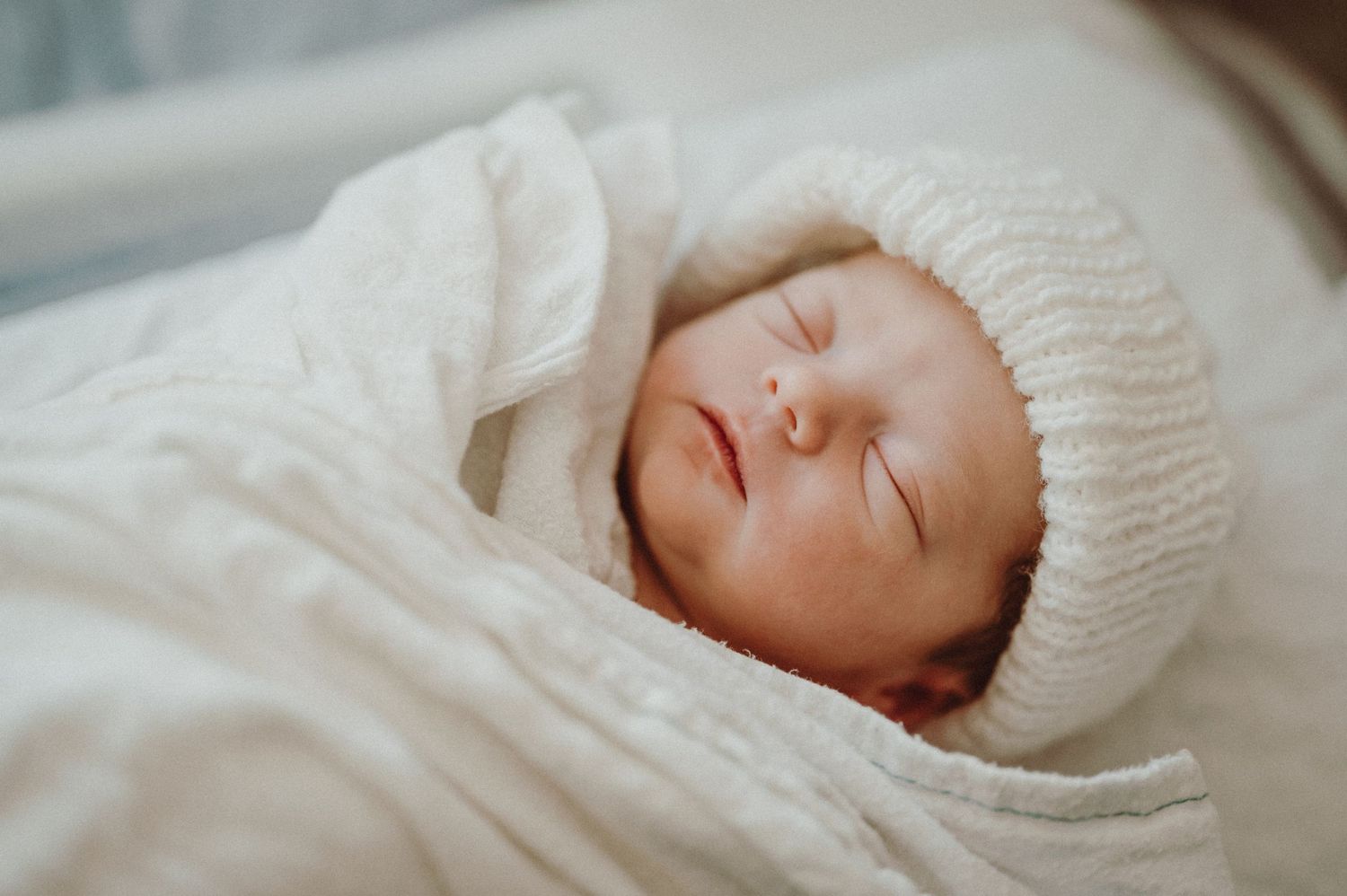 A sleeping newborn wearing a white knit hat rests peacefully on soft bedding.