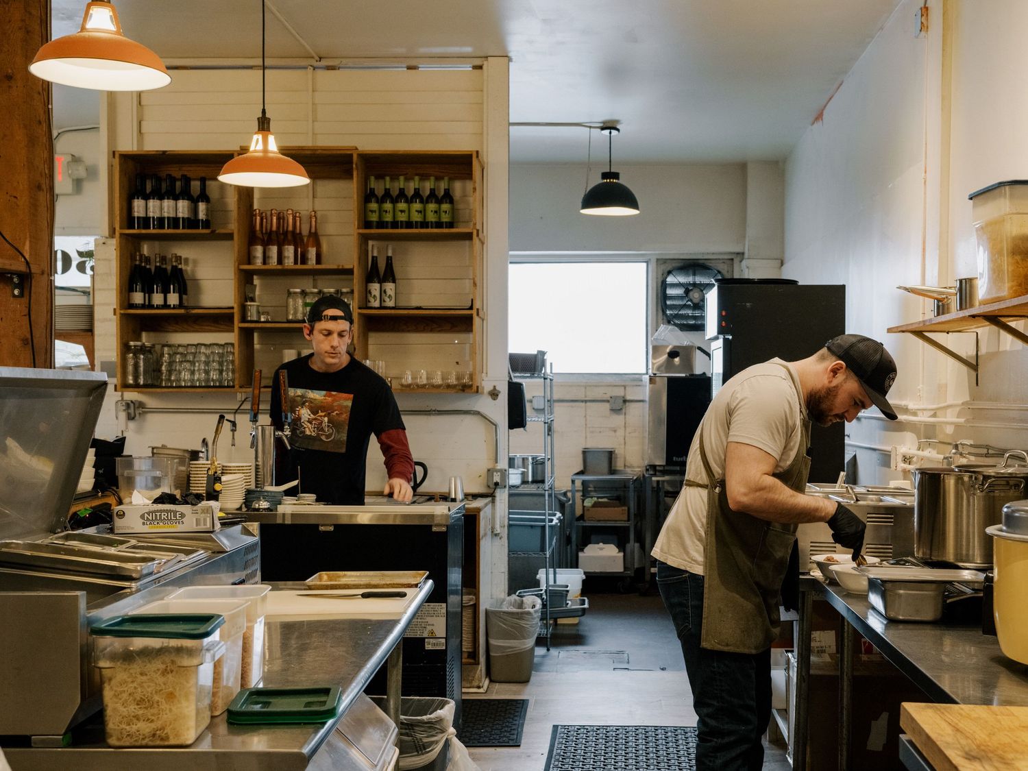 Restaurant kitchen workers preparing food in a commercial kitchen with hanging pendant lights and wooden shelving.