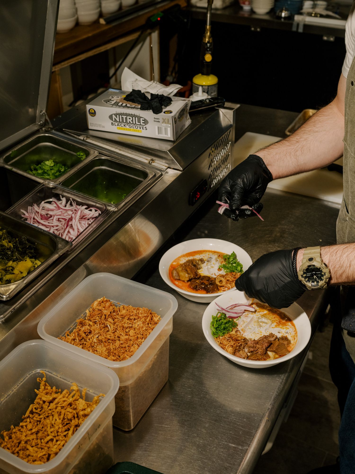 Restaurant kitchen prep station with various toppings and ingredients in metal containers.