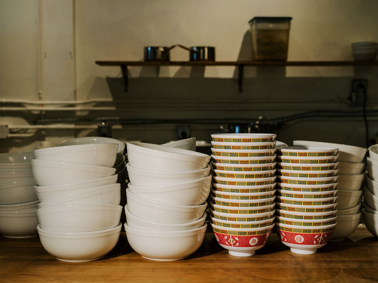 Stacks of white ceramic bowls and striped disposable cups arranged on a wooden shelf in a restaurant setting.