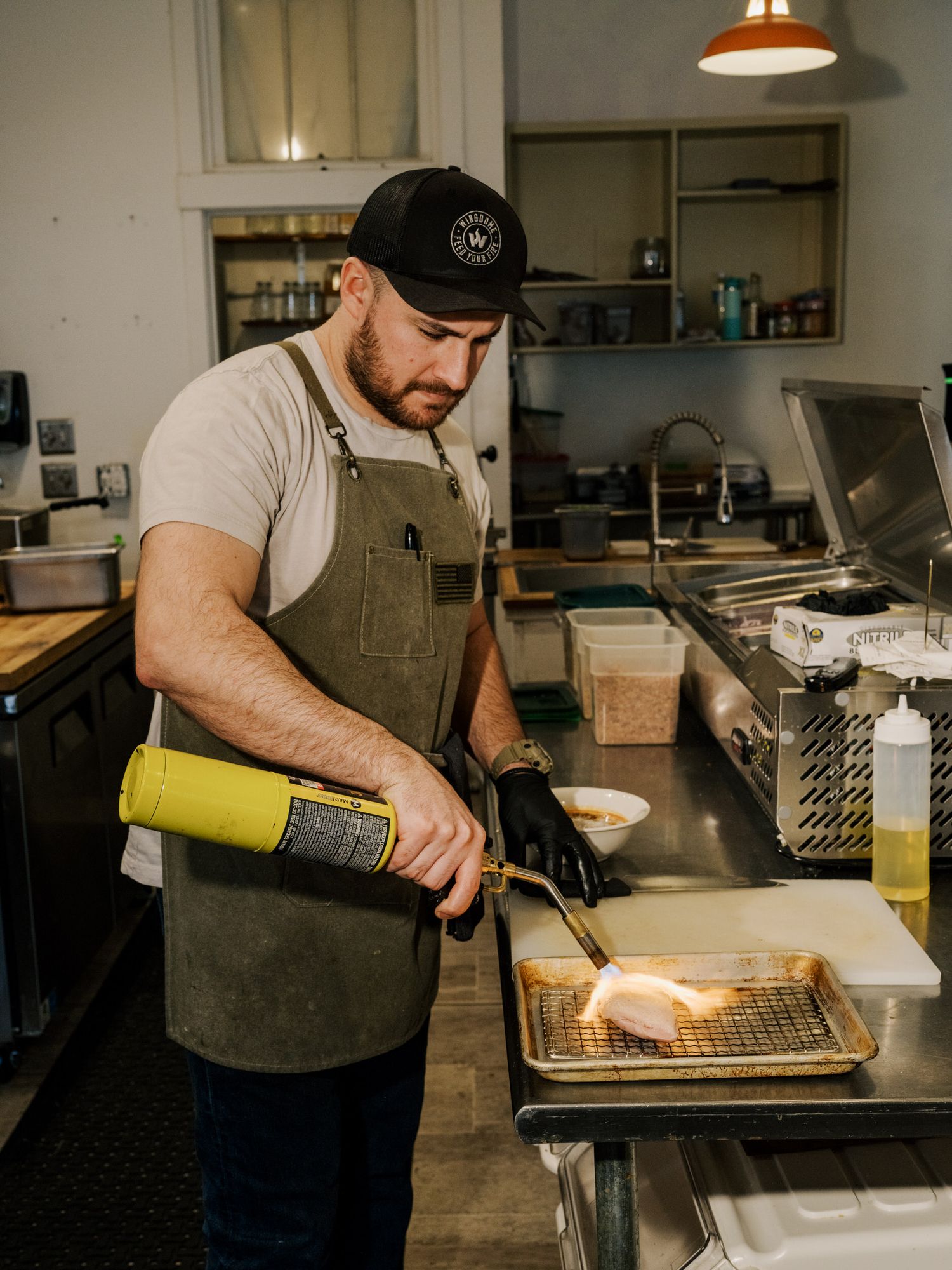 Chef using a culinary torch on food in a restaurant kitchen while wearing brown apron and black cap.